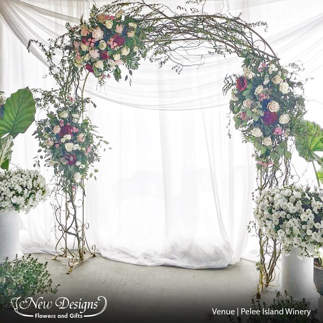 A Gold Curved Arch decorated with Full branches of curly willow and 3 installments of red and blush flowers, sits in front of a glowing white backdrop on an outdoor balcony.